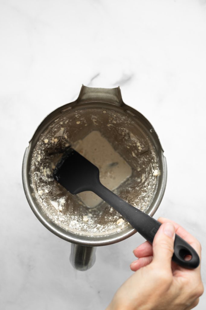Ingredients blended together in a blender container with a hand holding a black spatula scraping the sides.