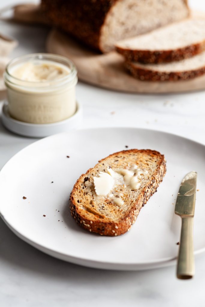 angled view of a buttered piece of toast with a knife on a white plate, loaf of bread and butter jar in the background.