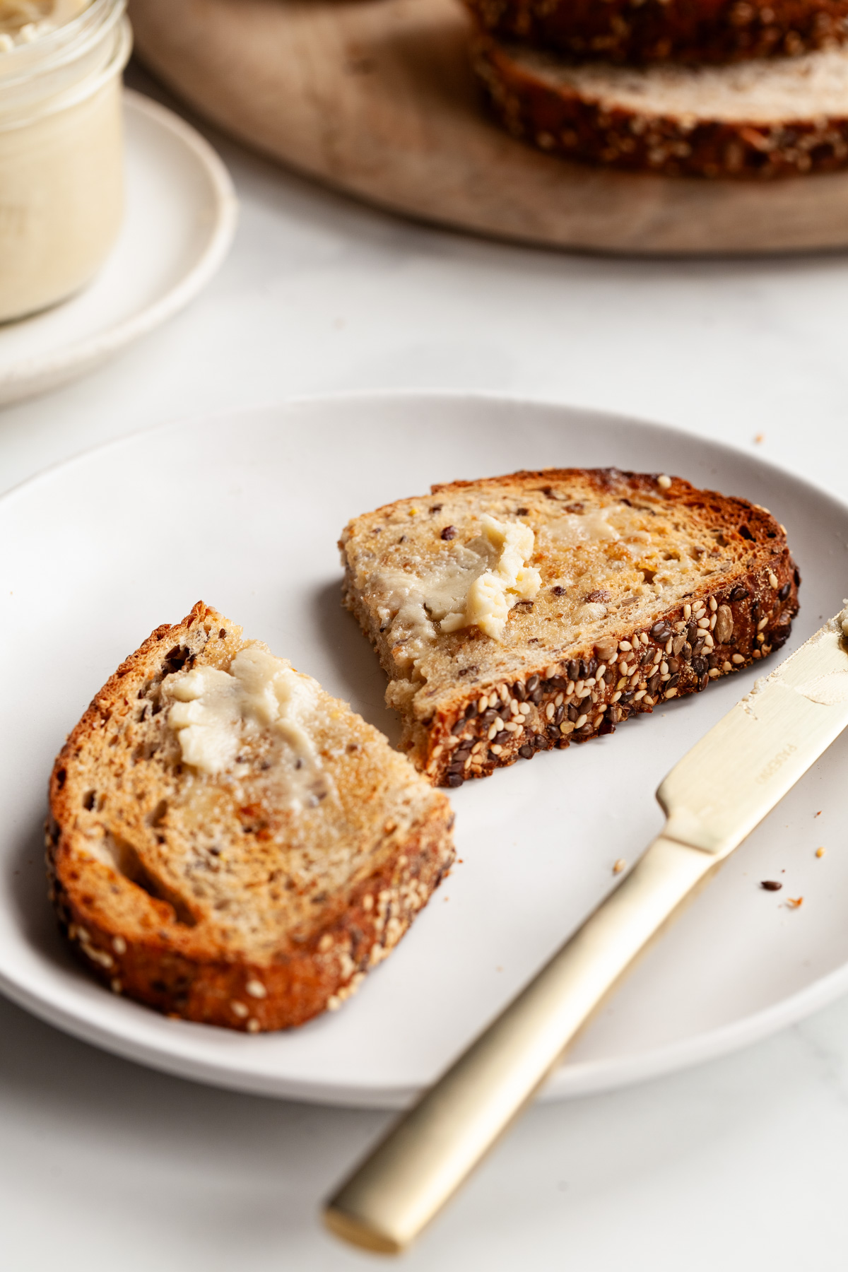 angled view of a buttered piece of toast with a knife on a white plate, loaf of bread and butter jar in the background.