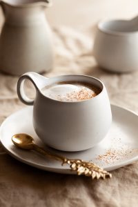 a mug filled with vegan pumpkin spice latte on a plate with a spoon on the side, a milk pitcher and mug are in the background.