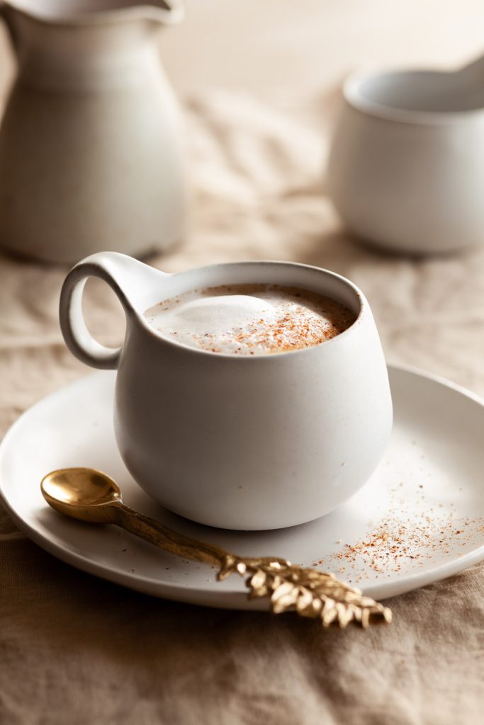 a mug filled with vegan pumpkin spice latte on a plate with a spoon on the side, a milk pitcher and mug are in the background.