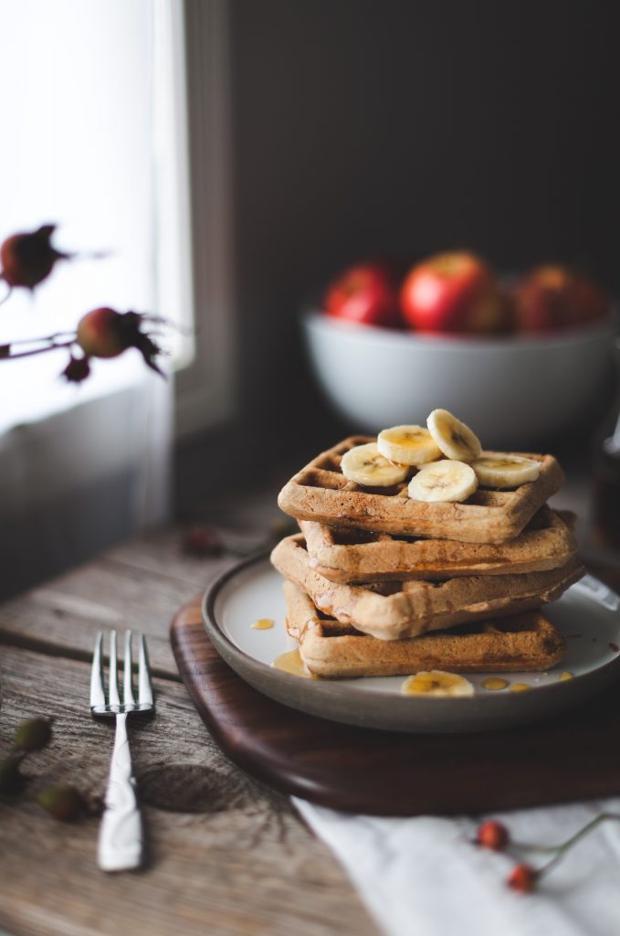 a head on image of a stack of 4 waffles on a white plate on a wooden surface with a bowl of apples in the background.