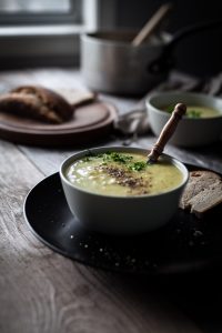 a bowl of leek potato soup with a spoon and slice of bread sitting near a window.