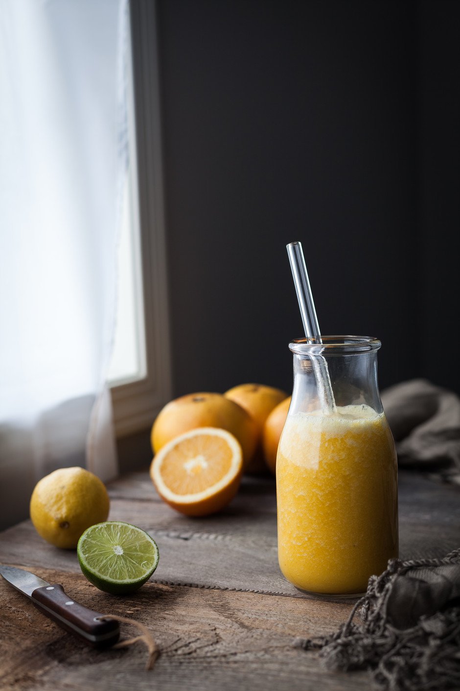 a glass of orange juice with a straw next to w indow with citrus.