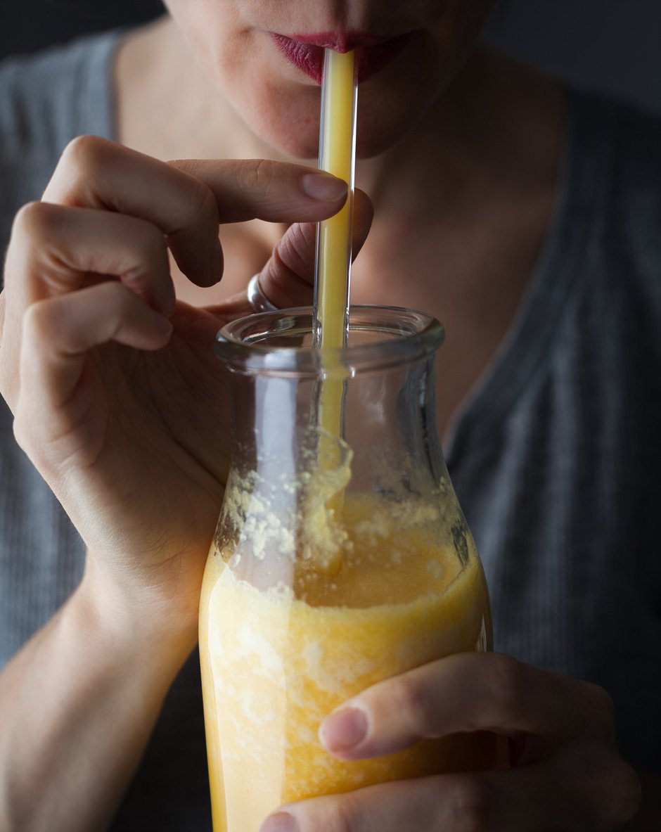 a close up of a person wearing a grey shirt sipping on a glass jar of citrus ginger juice with a glass straw in it.