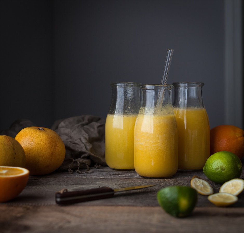three glass jars of juice sitting on a wooden table with citrus fruits and a knife.