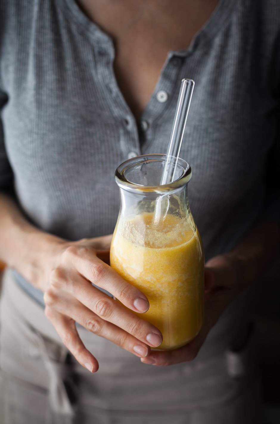 a close up of a person wearing a grey shirt holding a glass jar of citrus ginger juice with a glass straw in it.