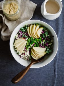 overhead of kale slaw with apples on a blue surface with a jar of parmesan and dressing.
