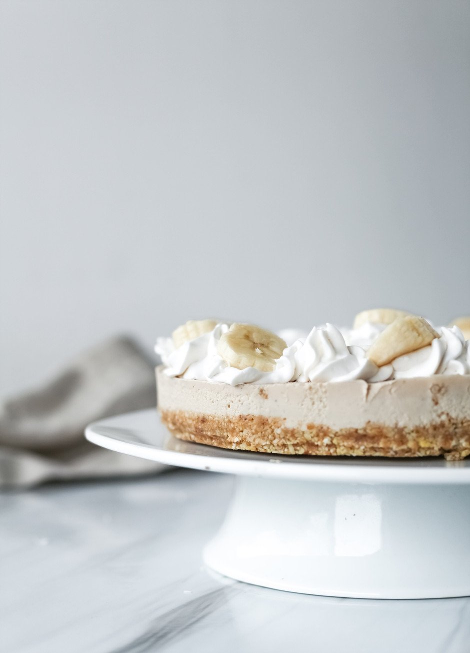 side view of a raw banana cream pie on a white cake stand.