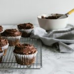 portrait of frosted chocolate cupcakes on a wire rack with a white bowl in the background on a marble surface.