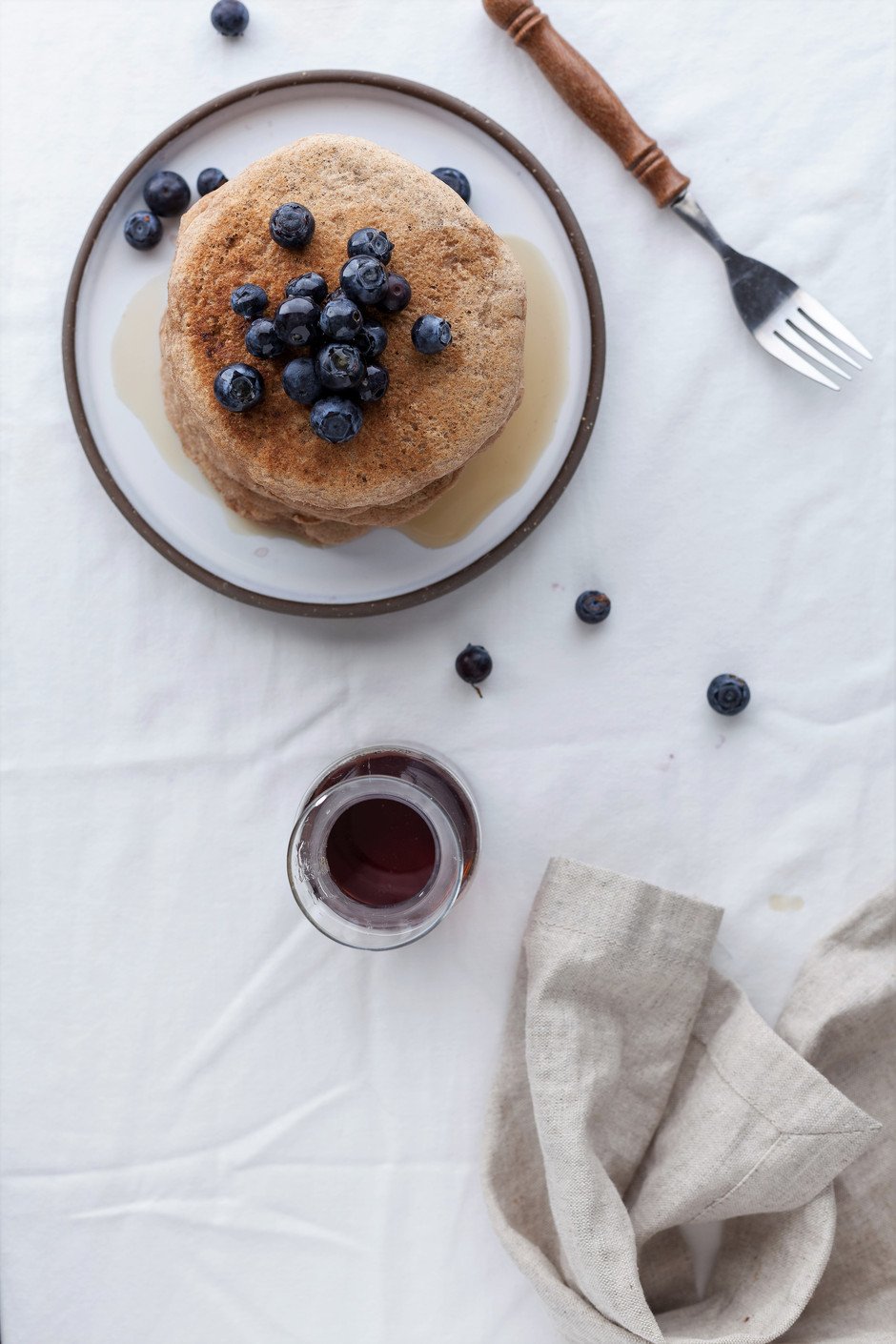 overhead image of vegan spelt pancakes with blueberries ontop sitting on a white linen tablecloth.