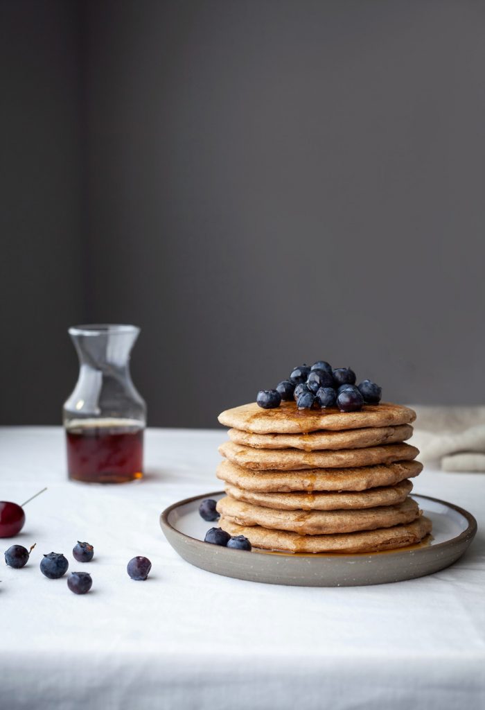 a head on image of a stack of 7 pancakes on a white plate on a white surface with a jar of syrup in the background.