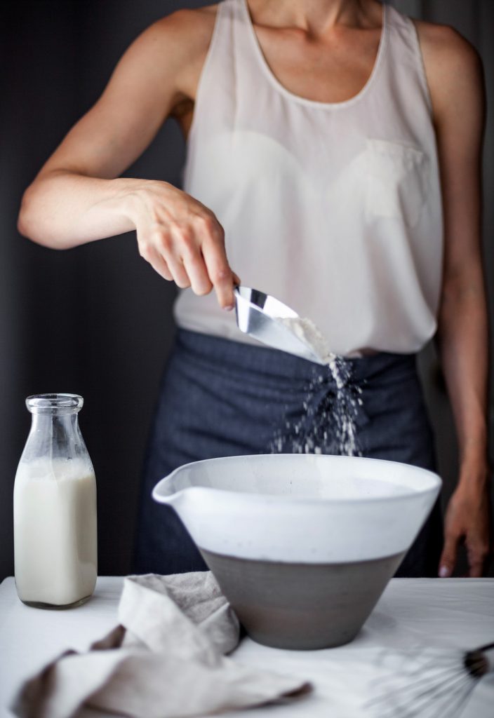 a baker sprinkling flour into a large ceramic mixing bowl with a jug of almond milk off to the side.