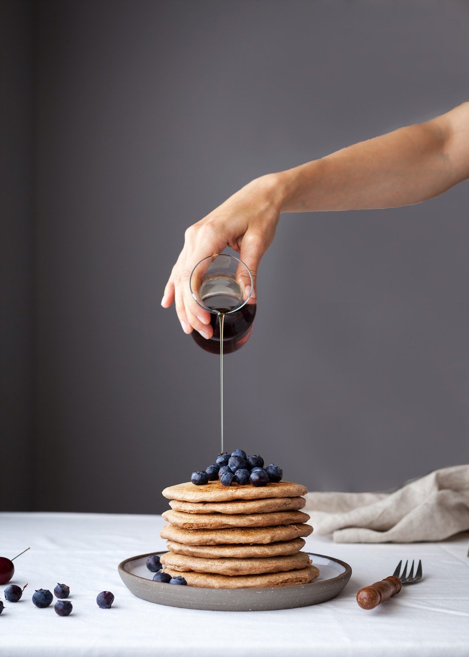head on image of a stack of pancakes with blueberries ontop with maple syrup being poured on top.