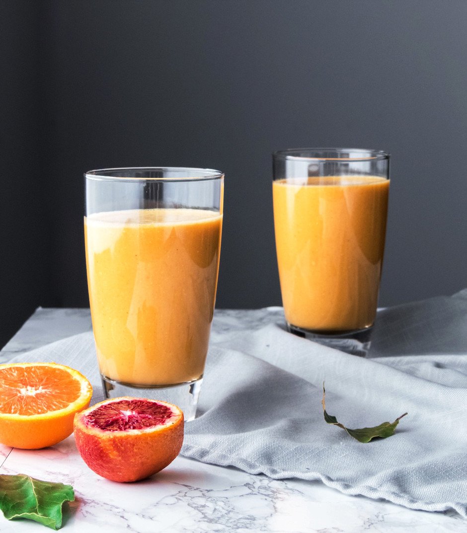 Straight on image of two glasses of citrus and root sunshine smoothie with oranges on the left and a blue linen. 