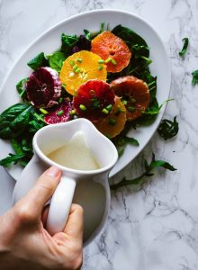 overhead view of a hand pouring a picther of dressing onto a salad with oranges.