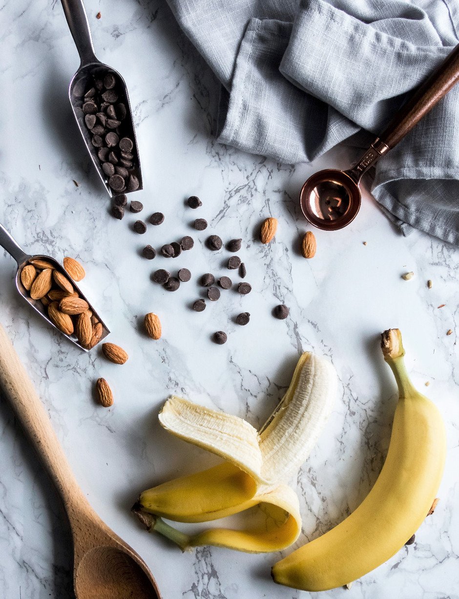 overhead image of bananas, almonds, chocolate chips and a wooden spoon on a marble surface.
