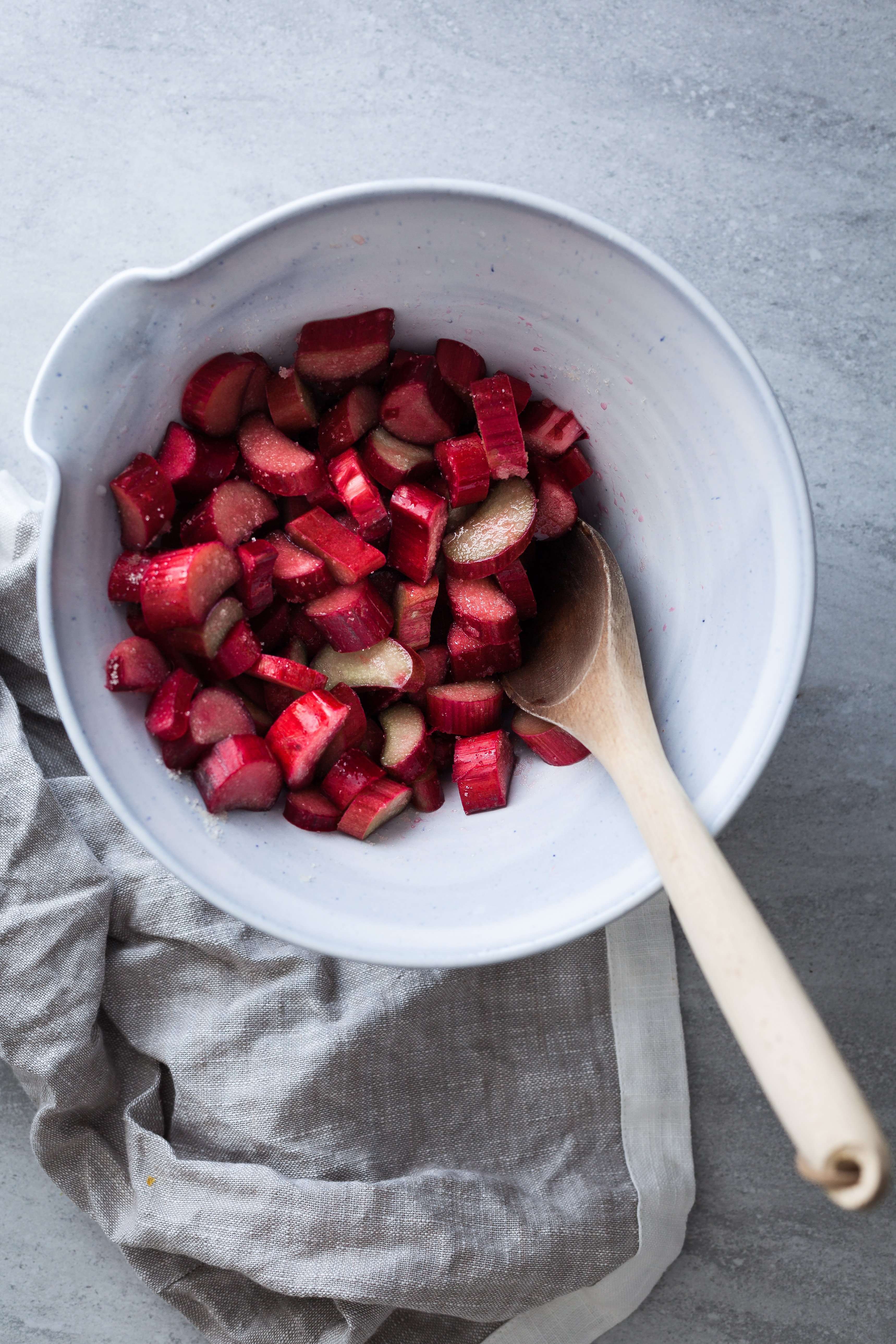 overhead image of chopped rhubarb in a white bowl with a wooden spoon and grey linen.