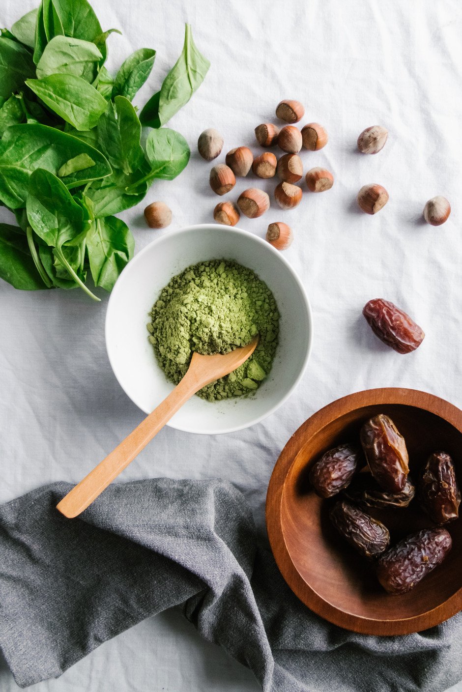 overhead of matcha tea with a wood spoon and a bowl of dates, spinach and hazelnuts on a white surface.