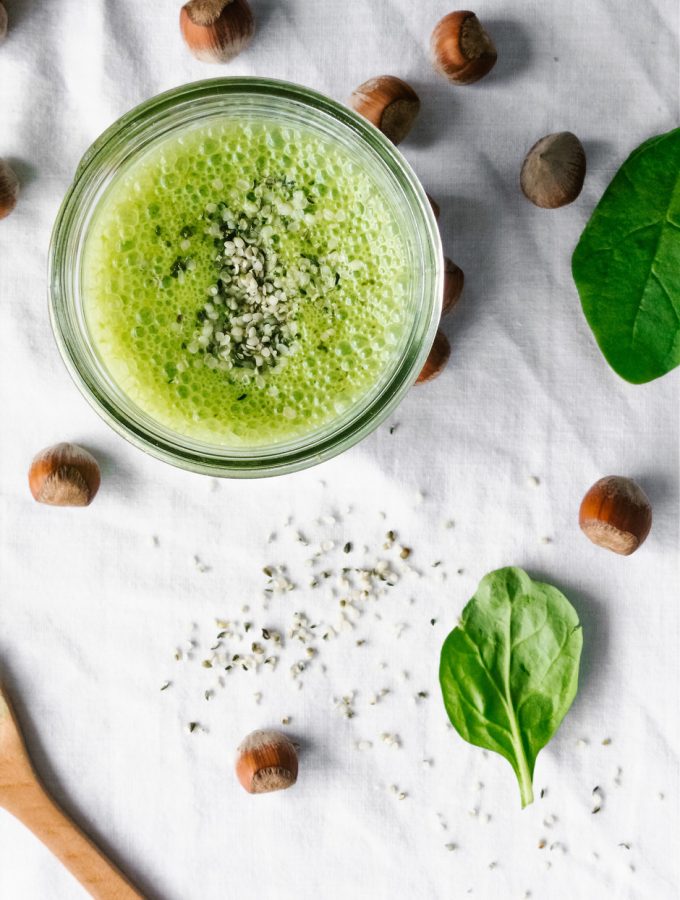 overhead of a green smoothie on a white surface with hazelnuts and spinach.