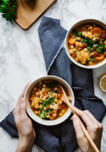two hands holding a bowl and spoon of soup on a blue linen with another bowl on the right..