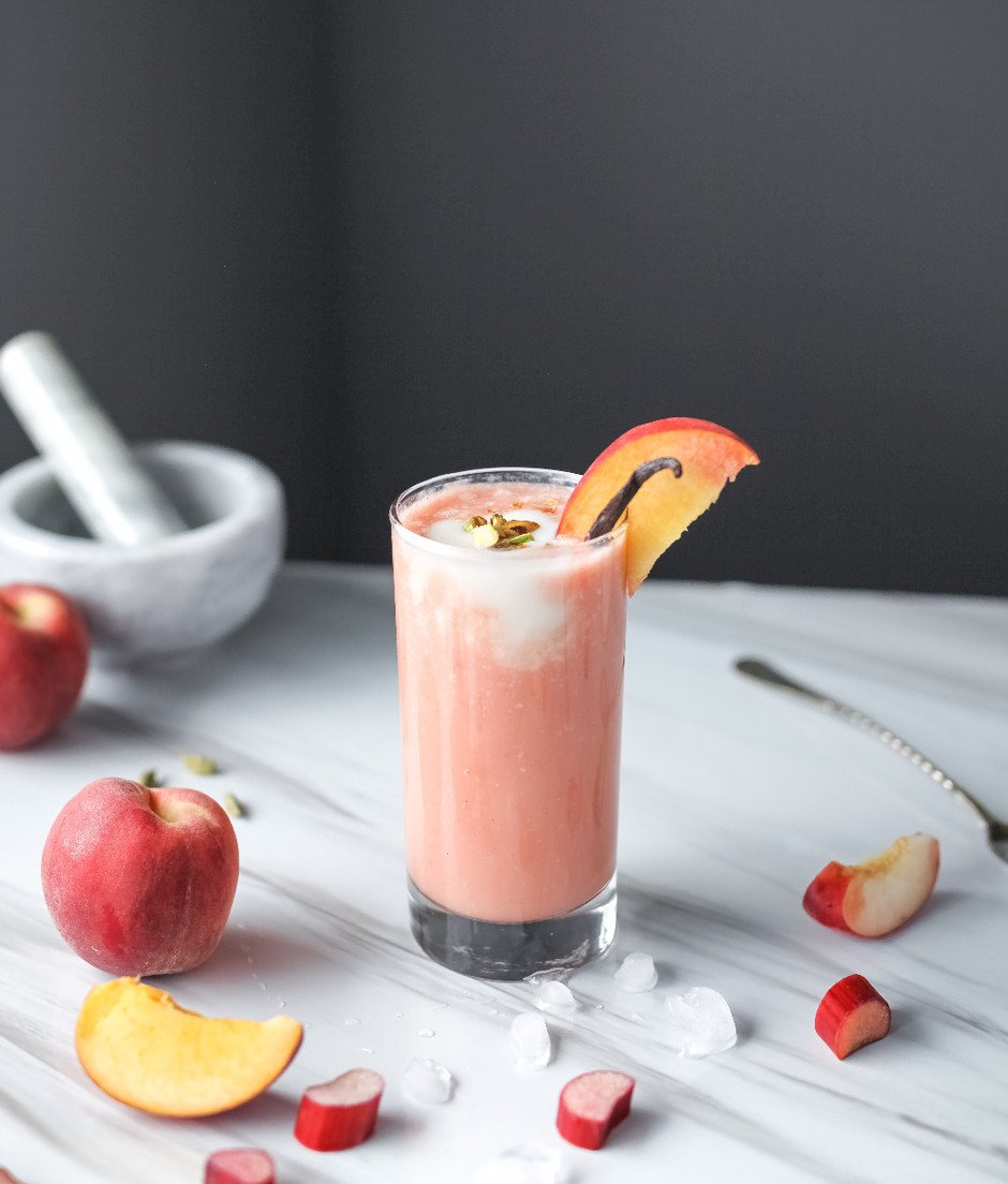A glass of peach lassi on a marble surface with peaches, ice and a mortar pestel.
