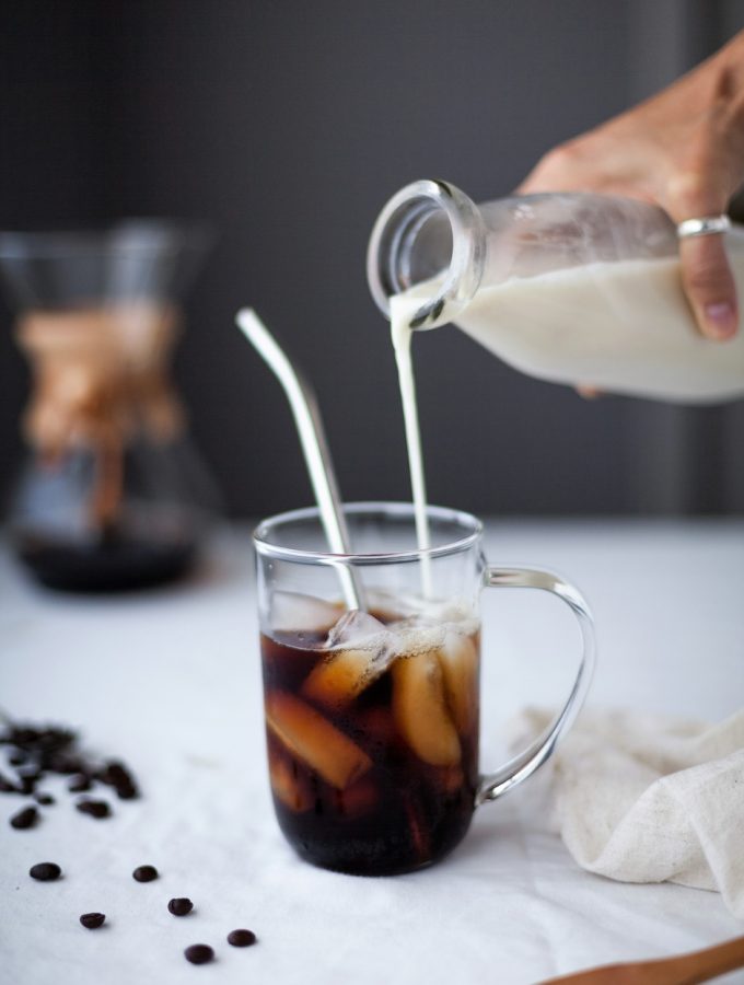 a person pouring milk into a glass of iced coffee with a straw.