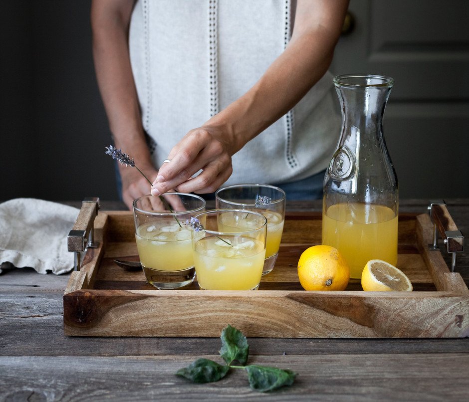 a person placing a lavender sprig in a glass of schorle on a tray with other glasses and lemons. 