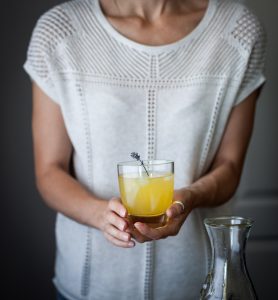 a person in a white shirt holding a glass of citrus schorle with a sprig of lavender.
