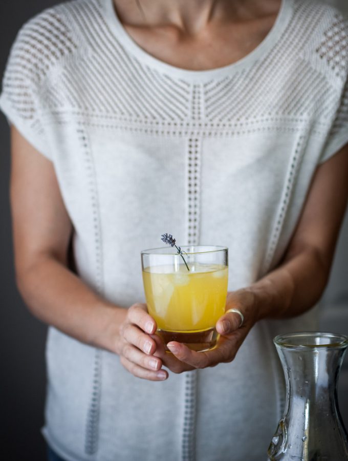 a person in a white shirt holding a glass of citrus schorle with a sprig of lavender.