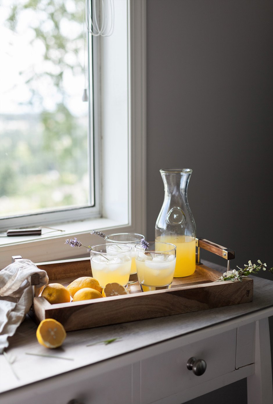 A bottle and glasses of schorle on a tray next to a window with lavender sprigs and lemons. 
