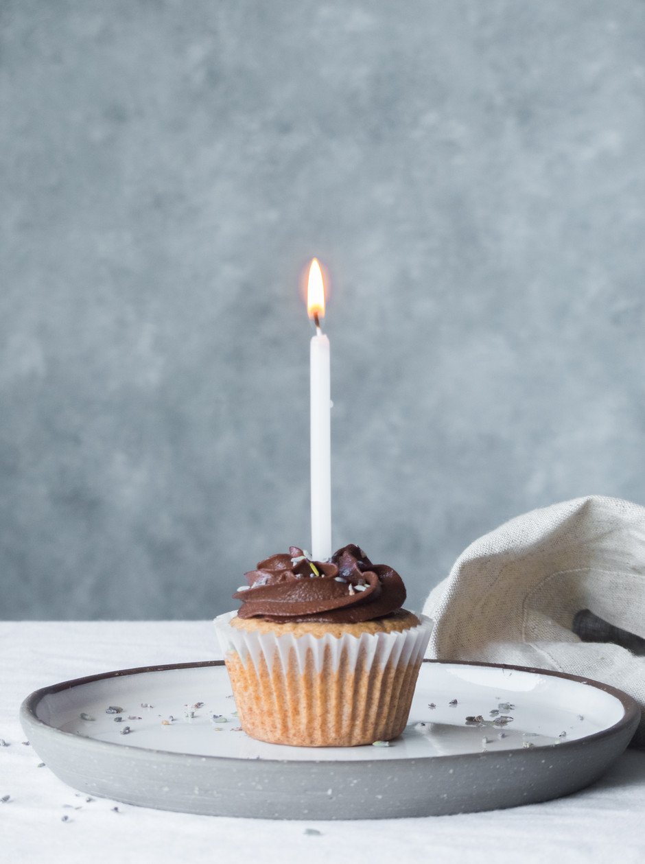 straight on view of a vanilla cupcake with chocolate frosting on a plate with one lit candle in it.