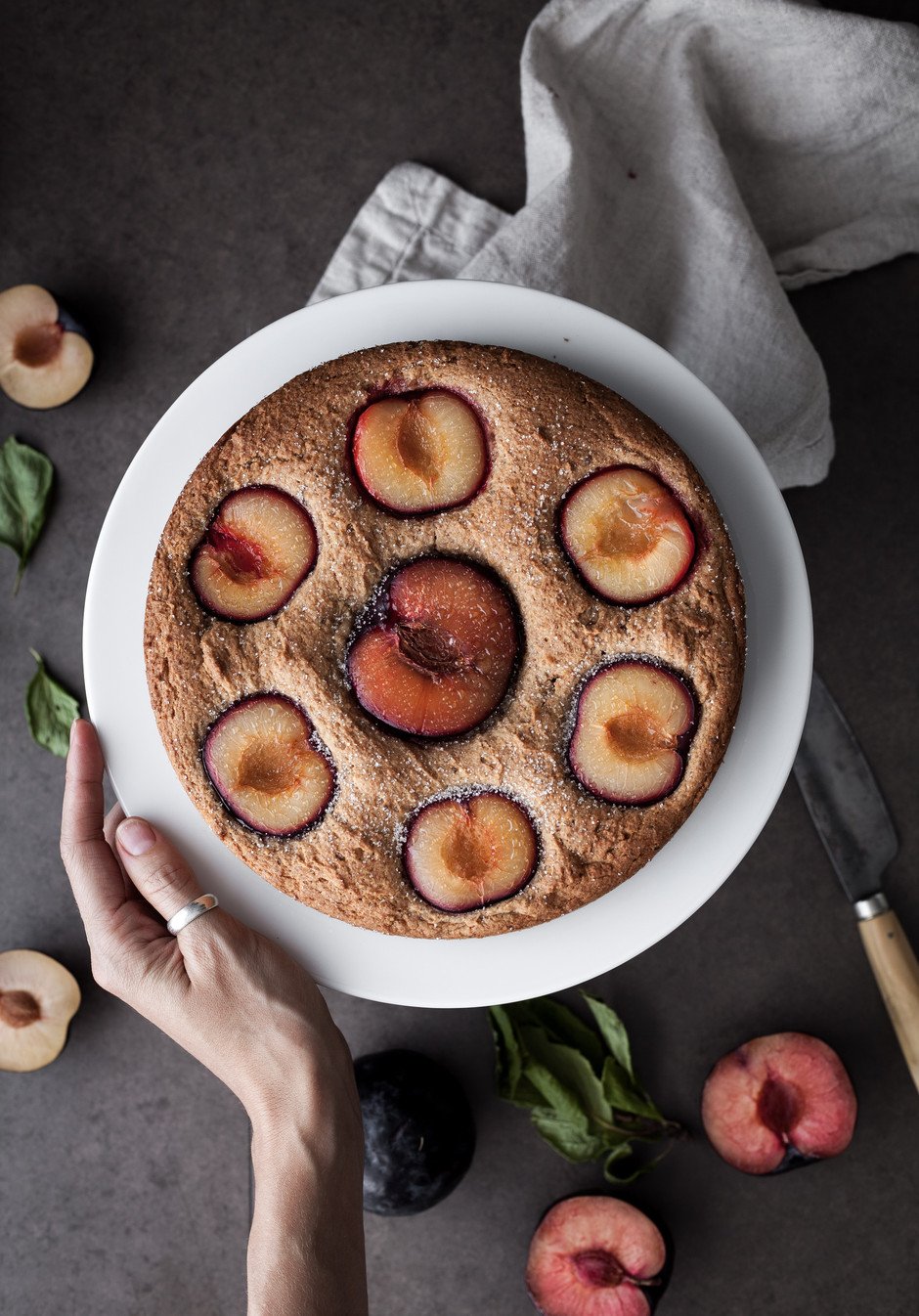 overhead view of a plum cake with a persons hand.