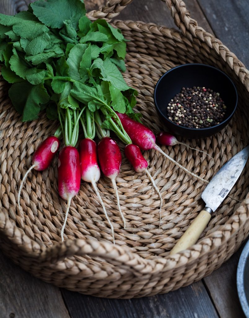 fresh radishes in a wicker basket with a knife and peppercorns on the right.