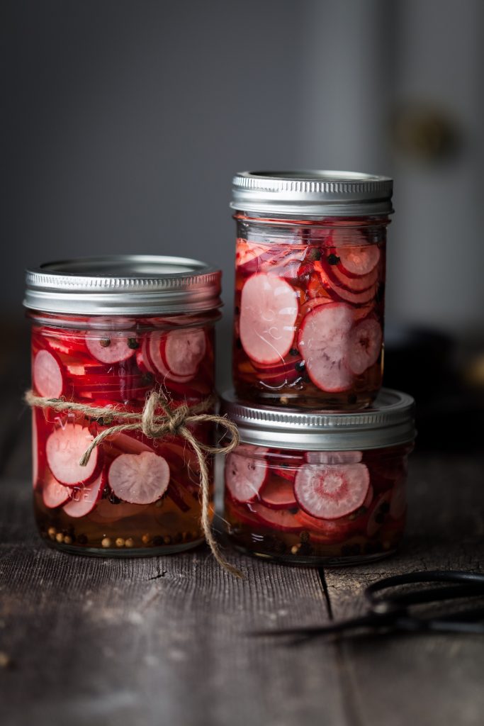 a portrait of three jars of pickled radishes sitting on a wood table.