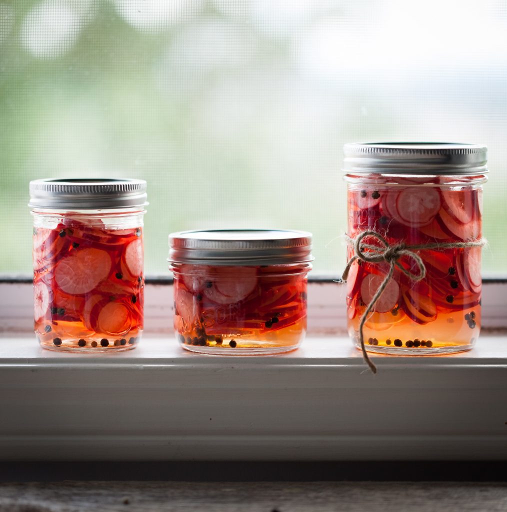 a portrait of three jars of pickled radishes sitting on a window sill.