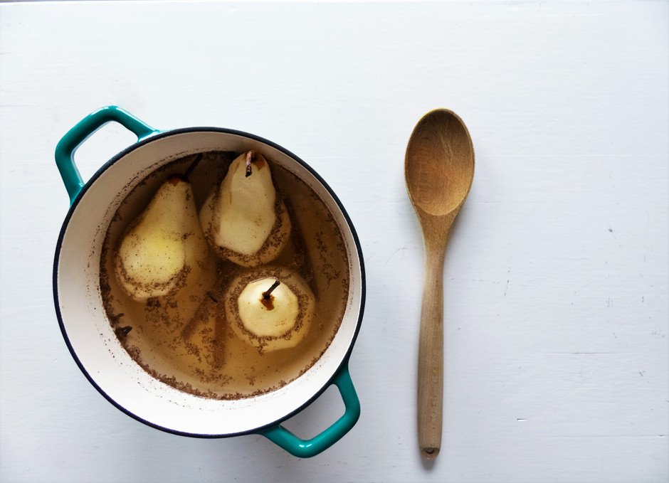 an overhead image of three pears being poached in a large dutch oven with a spoon off to the right.