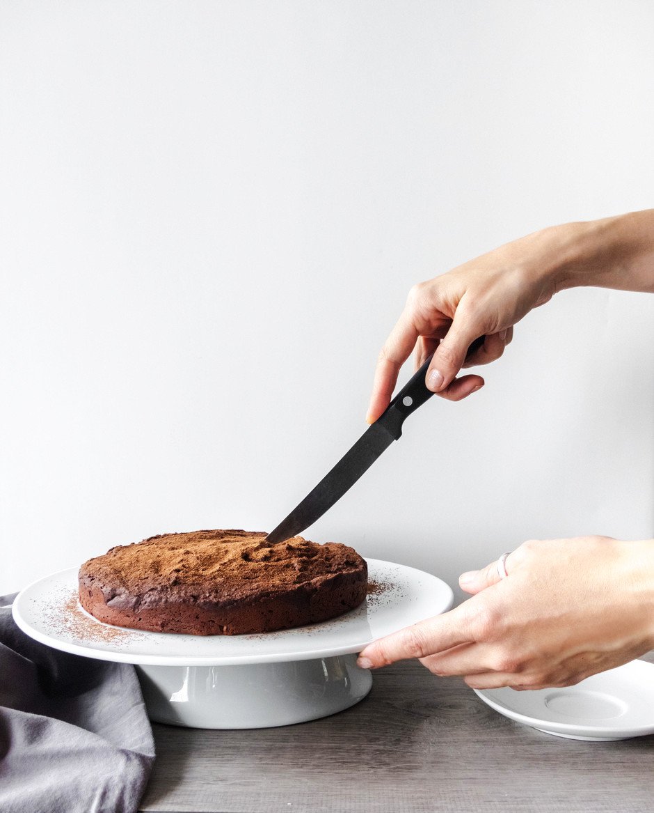 A person holding a knife about to cut into a chocolate torte sitting on a white cake stand against a white background.