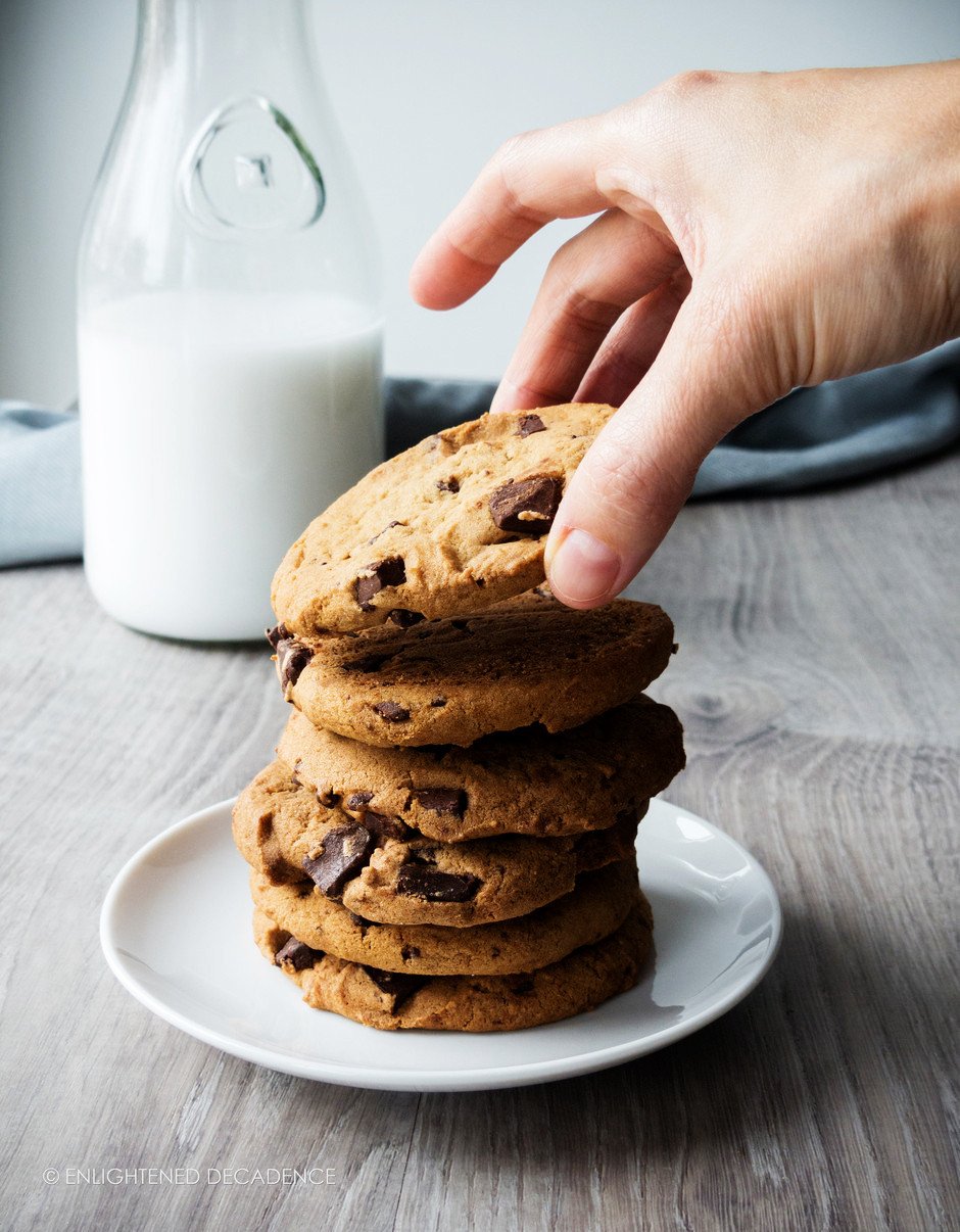 a head on image of a stack of chewy vegan chocolate chip cookies with a bottle of milk in the background and a hand reaching for a cookie.