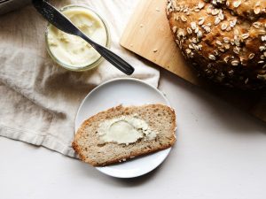 overhead of a slice of bread with butter, a loaf of bread, and jar of butter with a knife.
