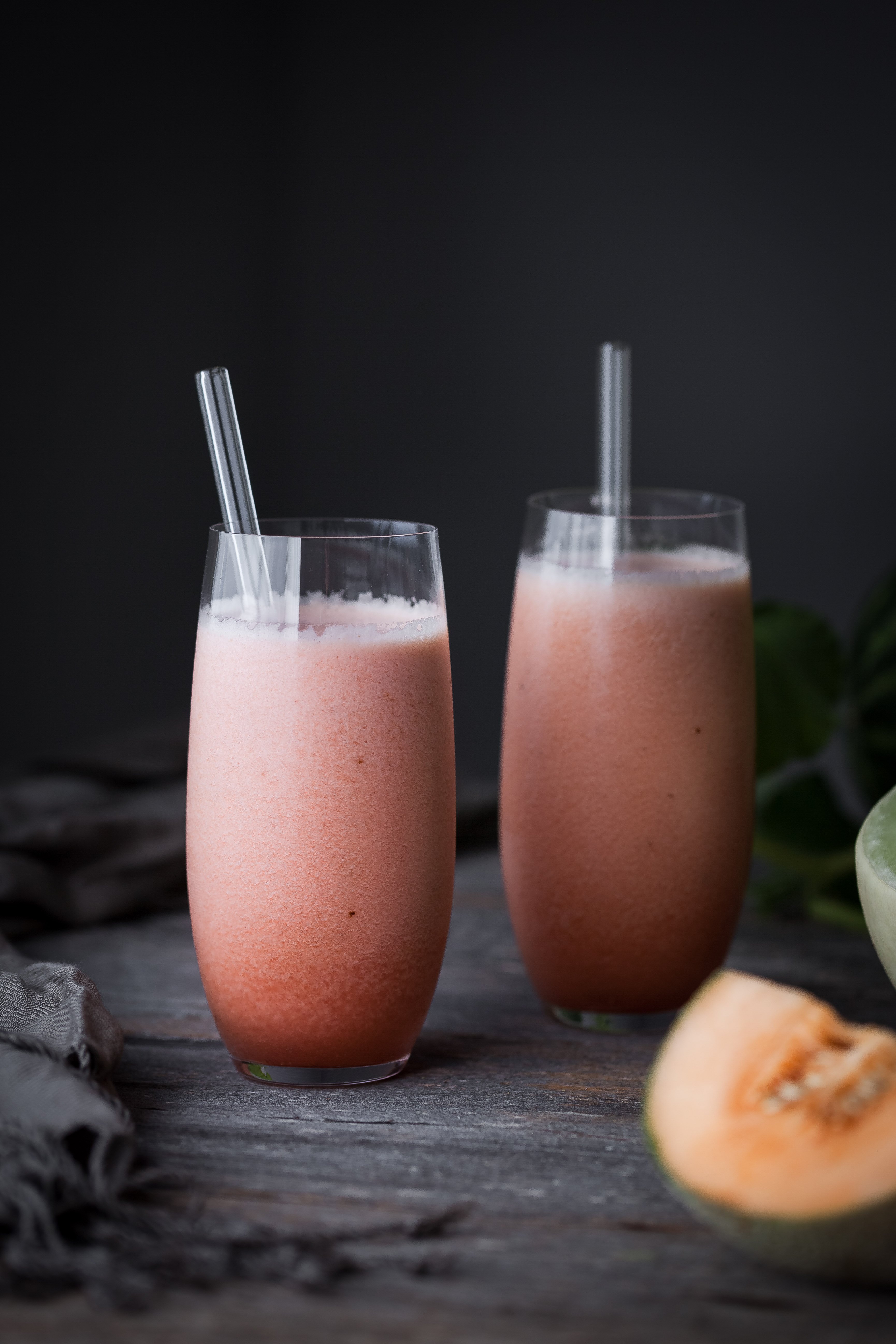 portrait view of two melon smoothies with straws on a wood table.