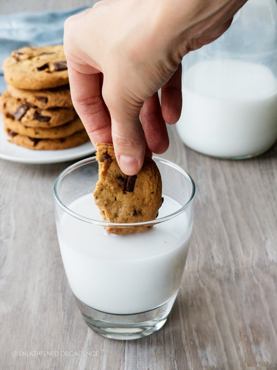 a hand dipping a piece of a chewy vegan chocolate chip cookie in a glass of milk.
