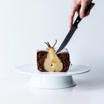 a hand holding a knife cutting into a pear cake on a cake stand.