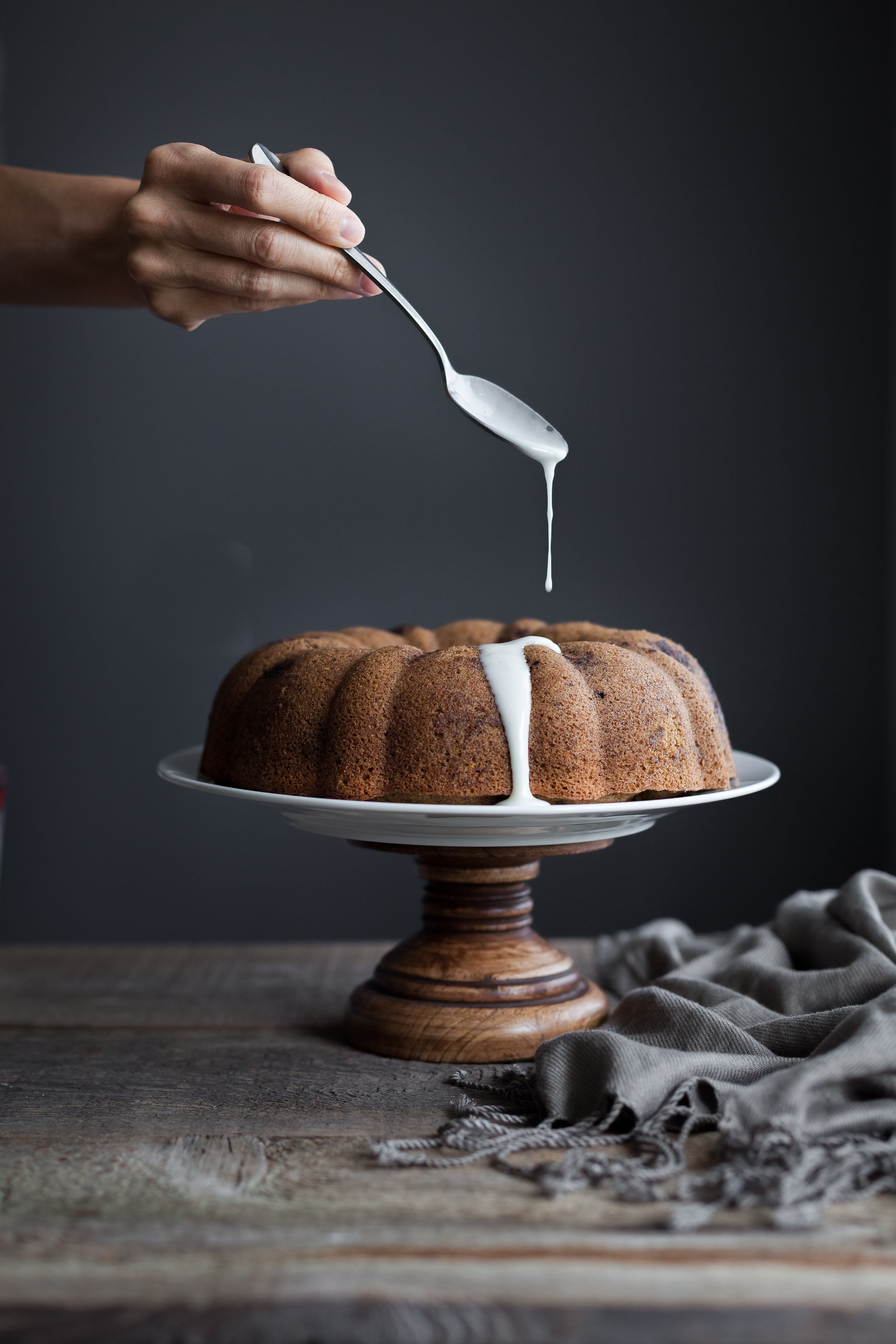 a hand holding a spoon drizzling glaze on bundt cake on a cake stand.