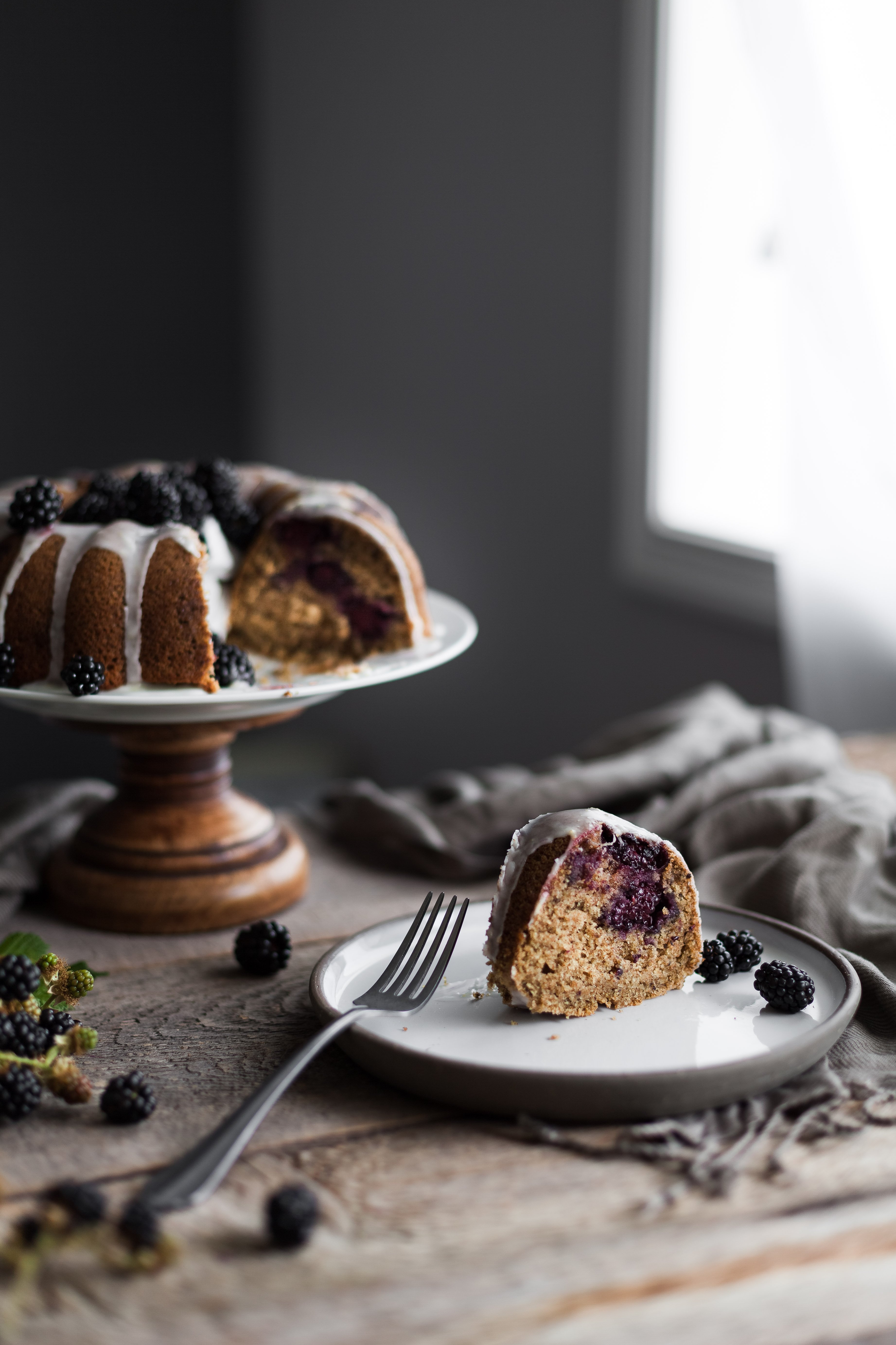 portrait of slice of blackberry cake on a plate with a cake on a cake stand in the background with a brown linen. 