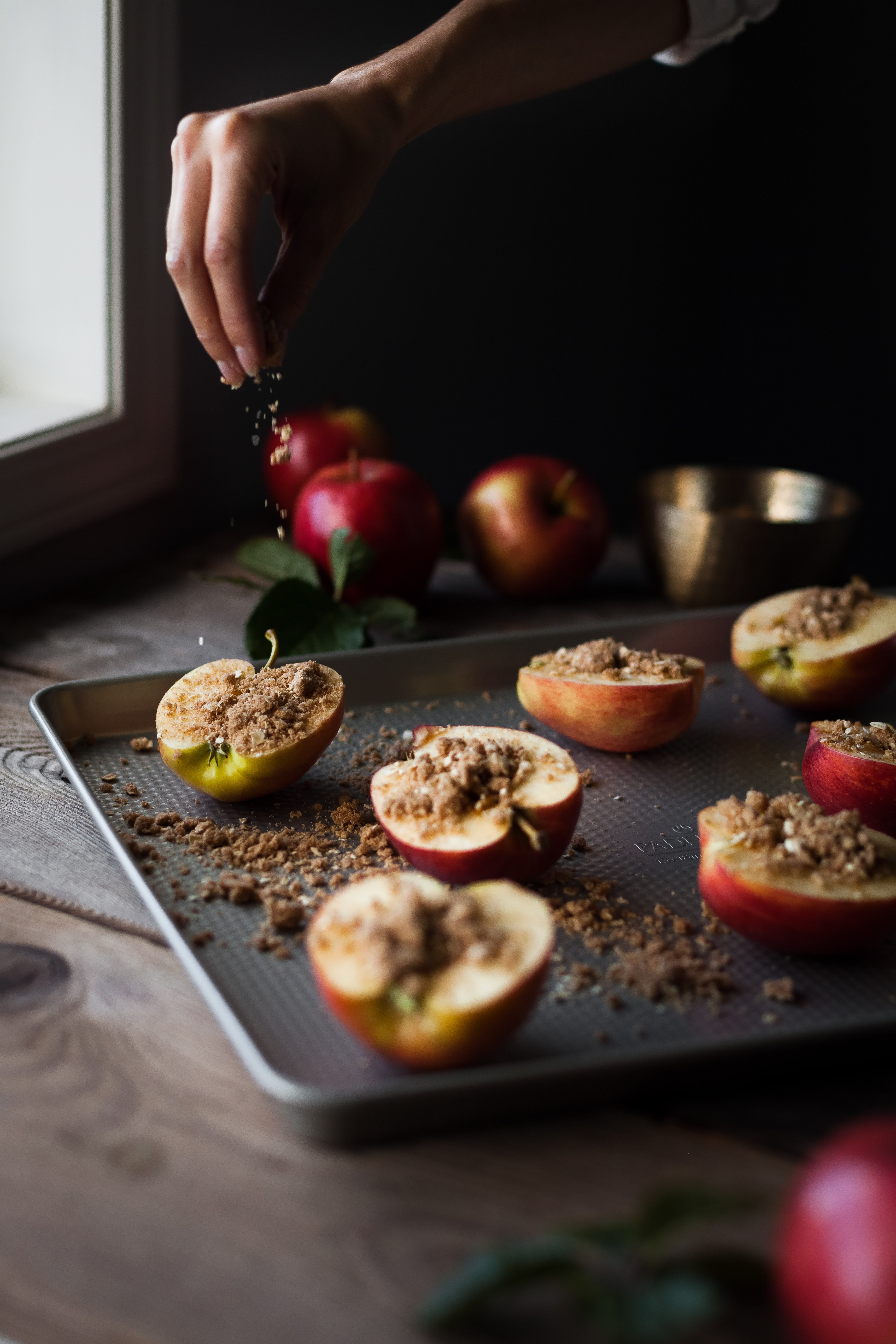 portrait of a hand sprinkling oat streusel on apples arranged on a baking sheet next to a window with apples in the background. 