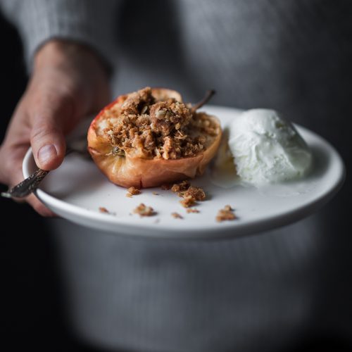 a person holding a white plate with a baked apple and ice cream on it.