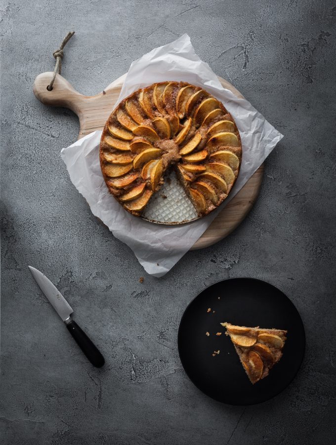 an overhead image of a german apple cake with a slice on a black plate and aknife off to the side.