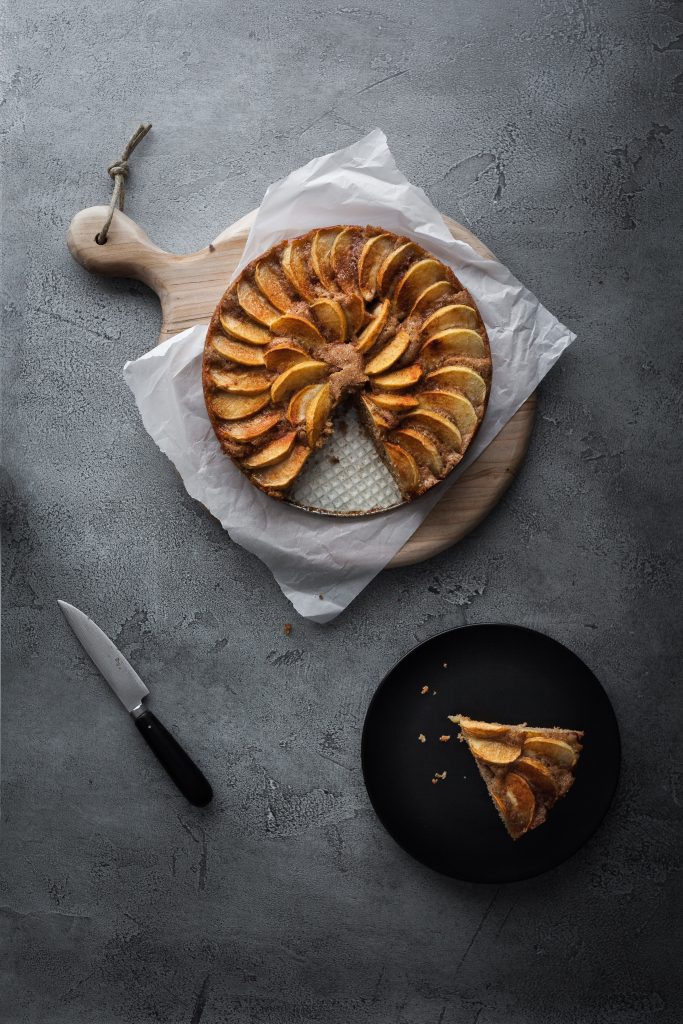 an overhead image of a german apple cake with a slice on a black plate and a knife off to the side.