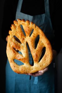a baker in a blue apron holding a fougasse bread.